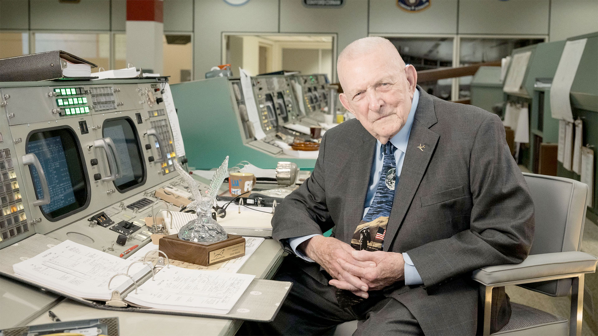 an older man wearing a jacket and tie sits at the restored console inside a historic mission control room